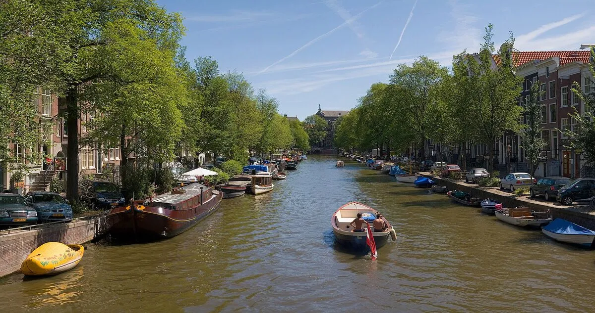 Amsterdamski kanał obsadzony drzewami, z zacumowanymi barkami i płynącą łodzią