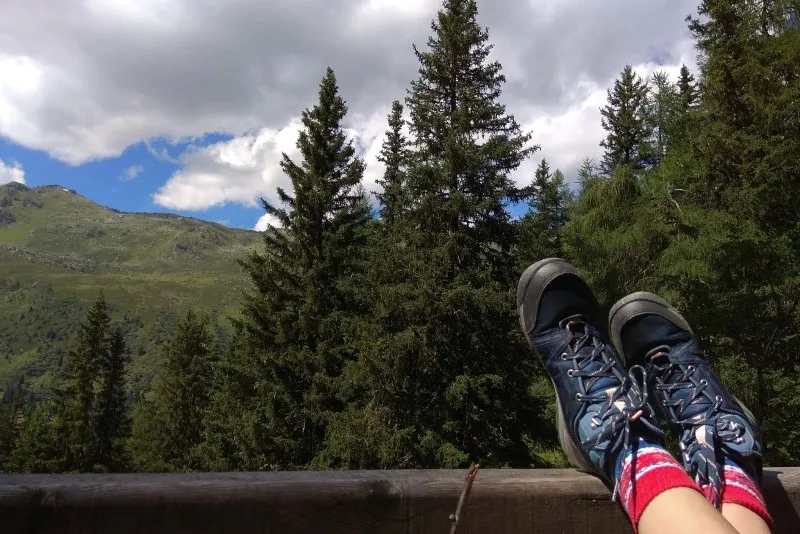 Nogi w butach trekkingowych oparte o drewnianą barierkę, w tle świerki i zbocze góry