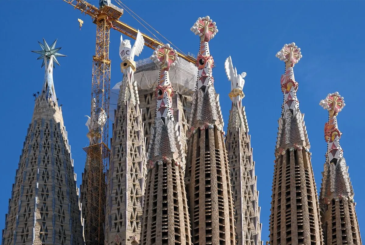 Zwieńczone mozaiką iglice wież Sagrada Família na tle błękitnego nieba, obok żuraw budowlany