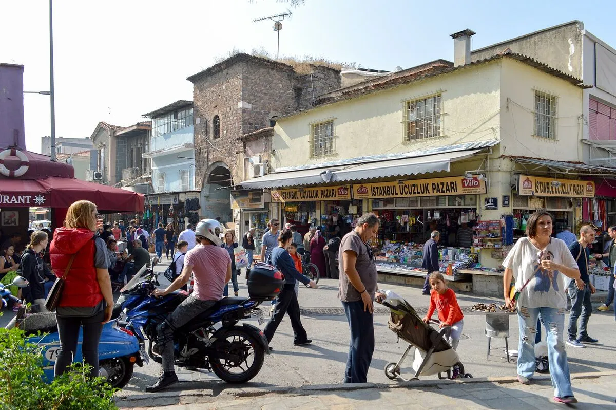 Przechodnie i sklepy na rynku w dzielnicy Kemeraltı w Izmirze, na pierwszym planie motocykl i kobieta z dzieckiem