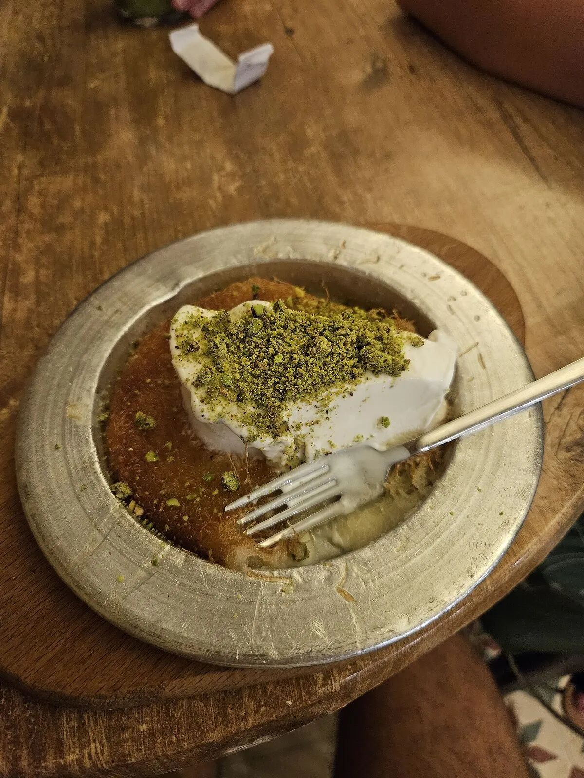 Kawałek kunafy z pistacjami na metalowym talerzu, z widelcem