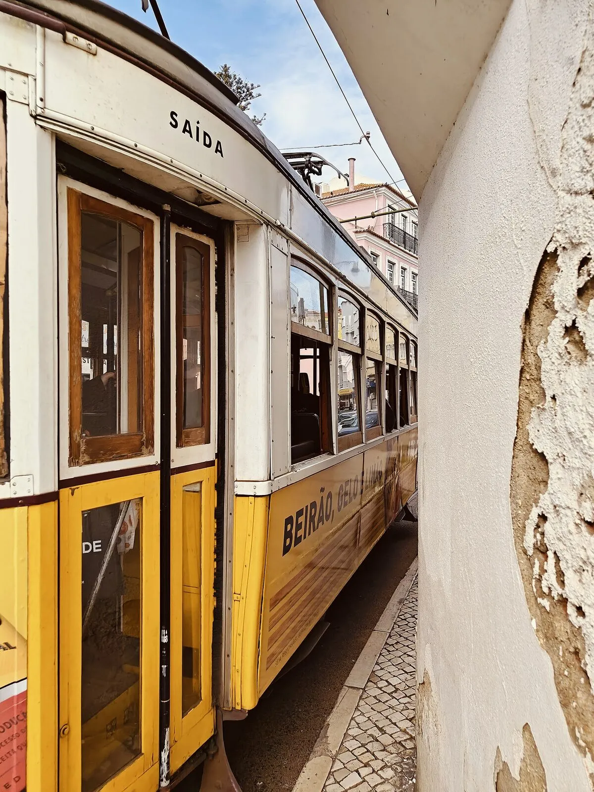 Żółto-biały tramwaj przeciskający się tuż przy ścianie kamienicy z odpadającym tynkiem
