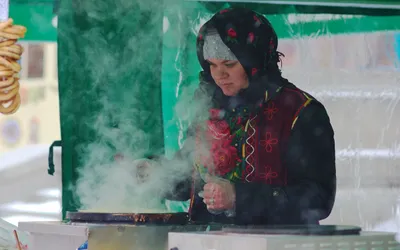 Kobieta w ludowym stroju smaży naleśniki na parującej płycie, obok wiszą obwarzanki