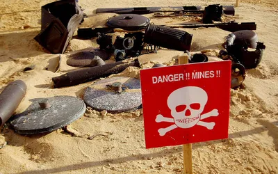 Czerwona tablica z czaszką i napisem „Danger!! Mines!!” obok rozbrojonych min na piasku (zdjęcie poglądowe z Libii)