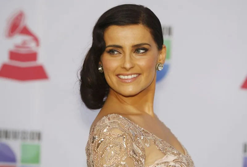 Uśmiechnięta Nelly Furtado w beżowej sukni na tle ścianki z logo Latin Grammy