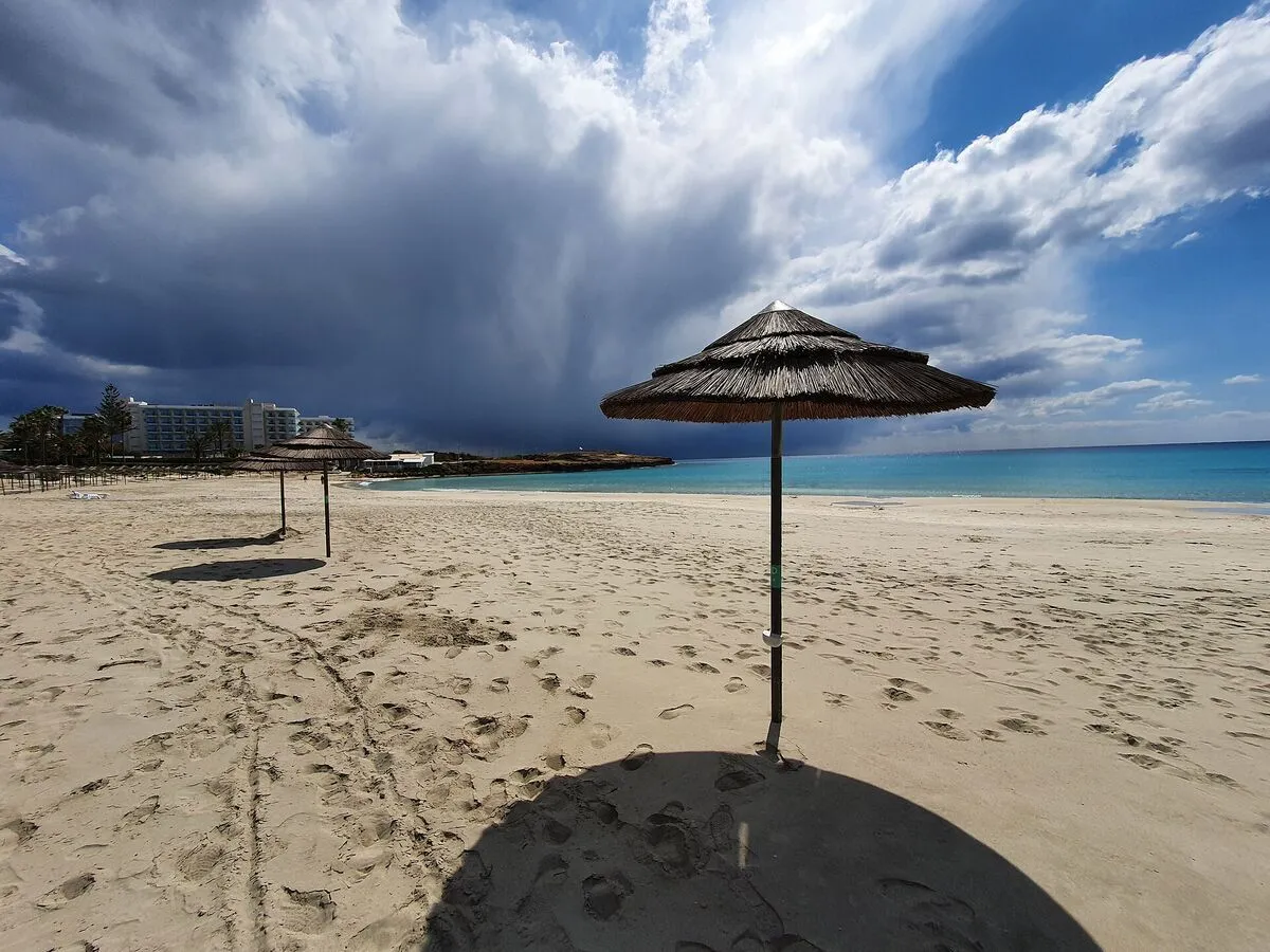 Trzcinowe parasole na pustej, piaszczystej plaży Nissi w Ajia Napie, w tle hotel i chmury nad turkusowym morzem