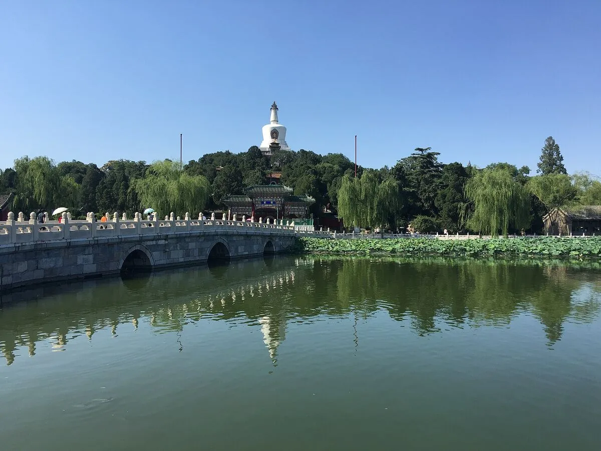 Biała dagoba na wzgórzu Wyspy Nefrytowej w parku Beihai, na pierwszym planie kamienny most i jezioro z liśćmi lotosu