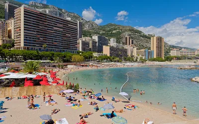 Plaża Larvotto z parasolami i kąpiącymi się ludźmi, w tle bloki i góry