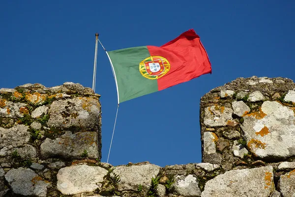 Flaga Portugalii Flaga Portugalii powiewająca nad kamiennymi blankami murów obronnych na tle błękitnego nieba