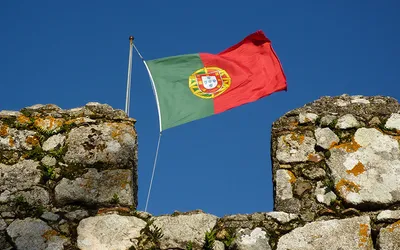 Flaga Portugalii powiewająca nad kamiennymi blankami murów obronnych na tle błękitnego nieba