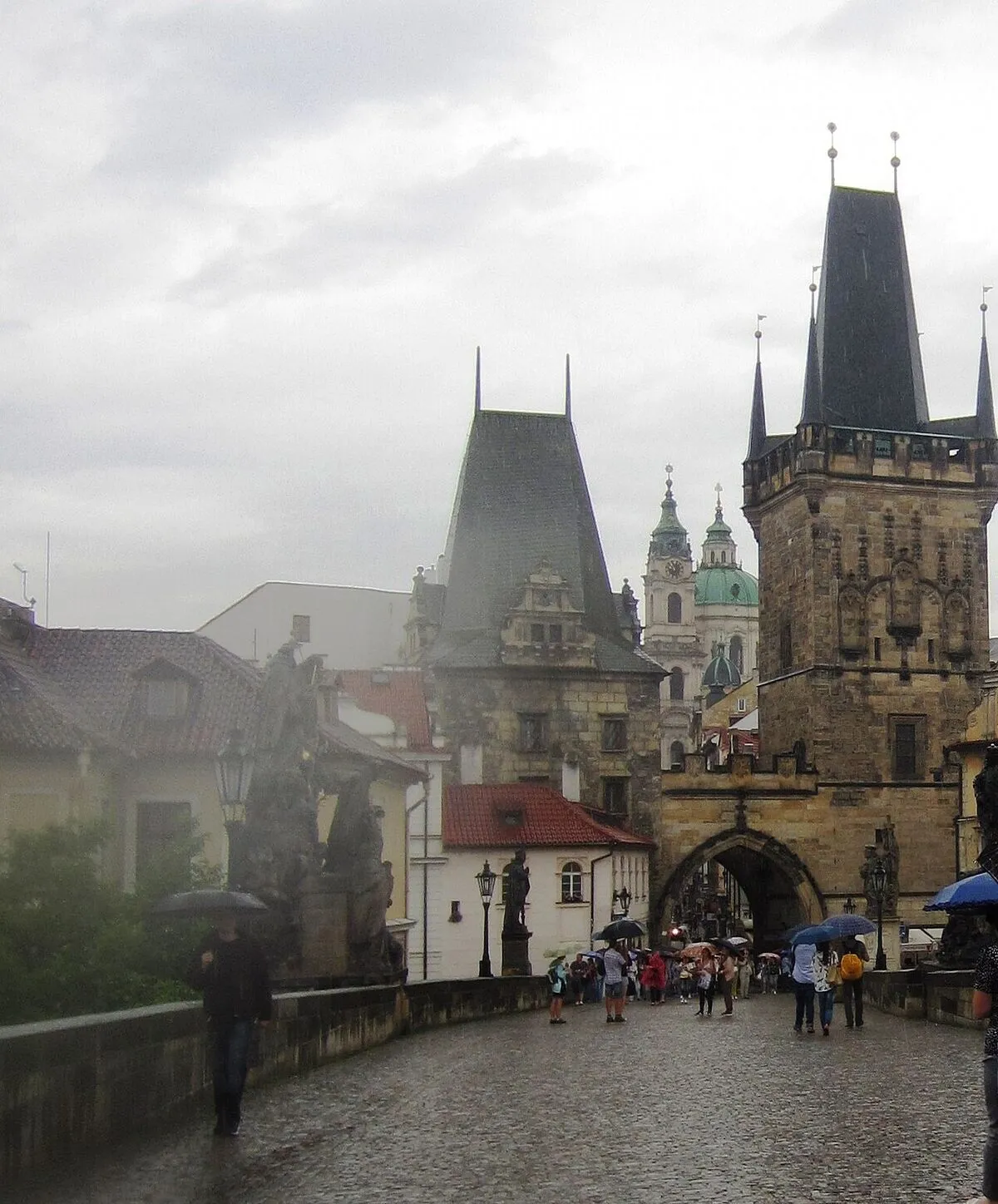 Mokry od deszczu bruk Mostu Karola, turyści z parasolami i wieże mostowe na Małej Stranie