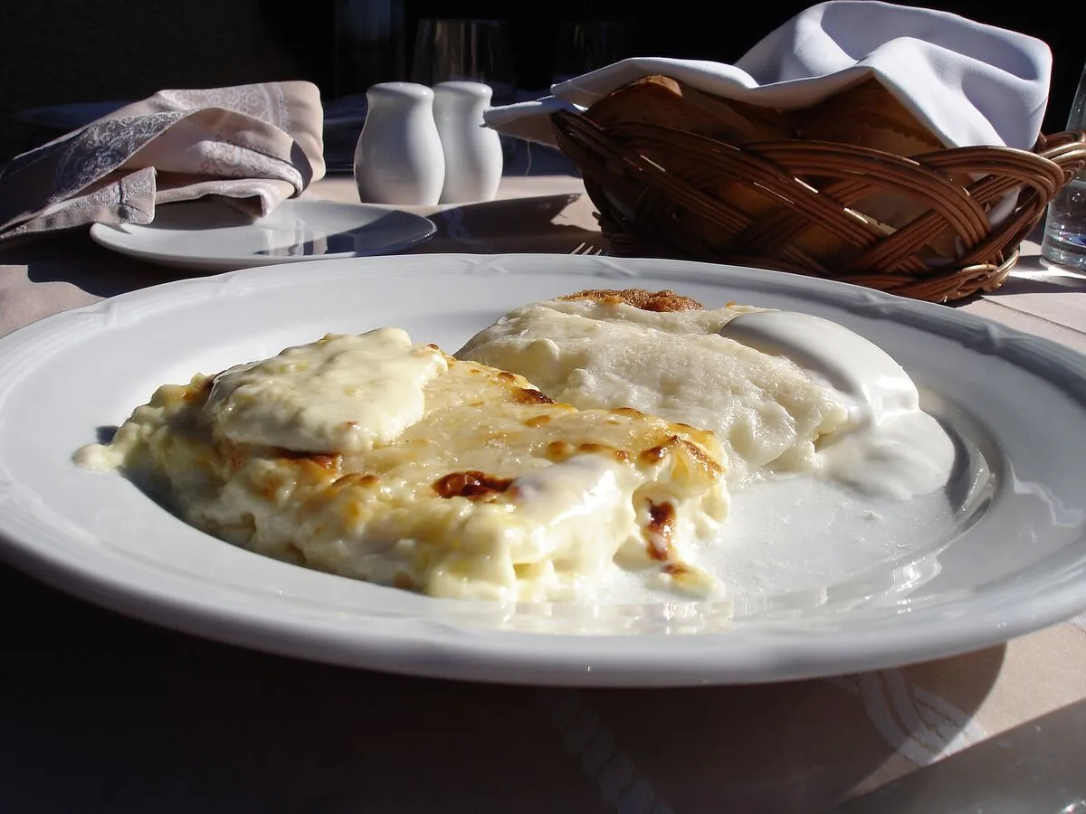 Porcja zapiekanych štrukli ze śmietaną na białym talerzu, w tle koszyk z pieczywem