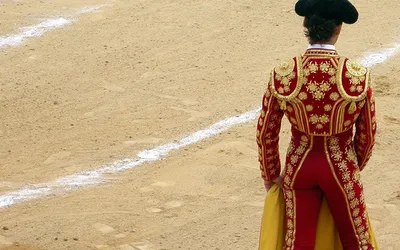 Torero w czerwono-złotym stroju z płachtą stoi tyłem na piaszczystej arenie