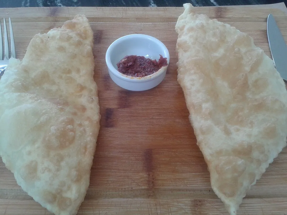 Dwa smażone çiğ börek na drewnianej desce, między nimi miseczka z czerwonym sosem