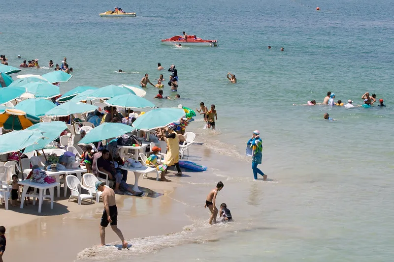 Zatłoczona plaża z turkusowymi parasolami, plastikowymi stolikami i kąpiącymi się ludźmi