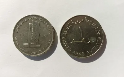 Dwie monety 1-dirhamowe na białym tle: okolicznościowa z wieżowcem banku i obiegowa z arabskim napisem i nazwą United Arab Emirates