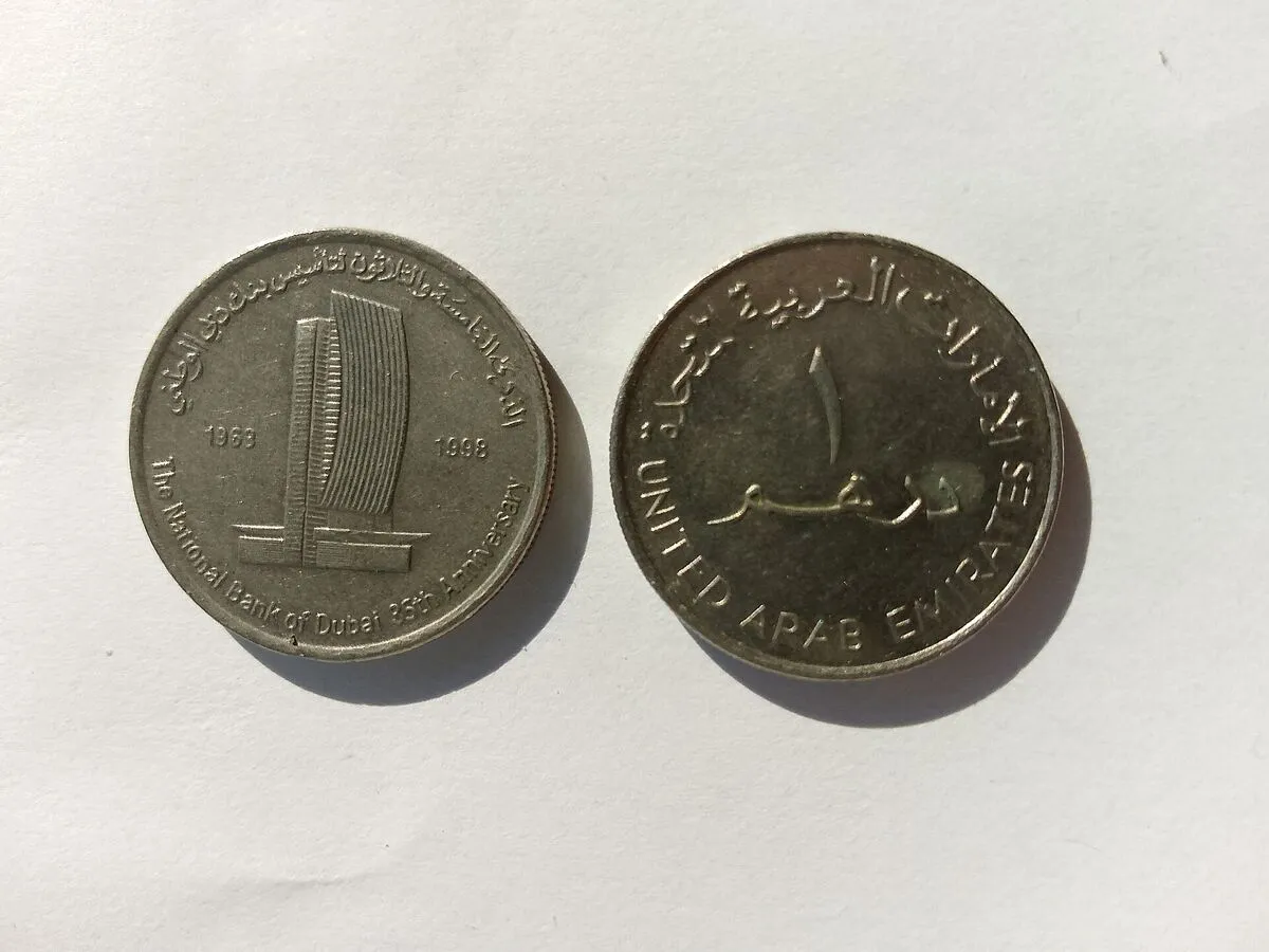 Dwie monety 1-dirhamowe na białym tle: okolicznościowa z wieżowcem banku i obiegowa z arabskim napisem i nazwą United Arab Emirates