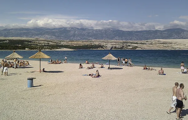 Plażowicze pod słomianymi parasolami na jasnej żwirowej plaży Zrće, w tle pasmo Velebitu