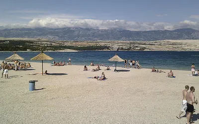 Plażowicze pod słomianymi parasolami na jasnej żwirowej plaży Zrće, w tle pasmo Velebitu